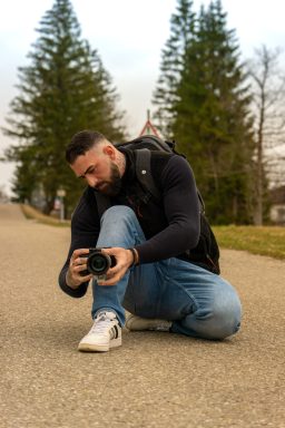 Mann in Jeans und Weste kniet auf der Straße und fotografiert mit einer Kamera.